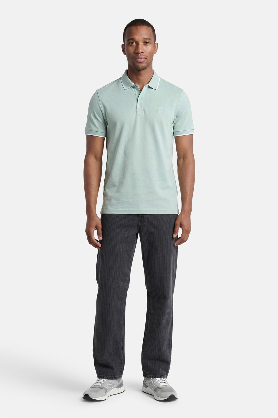 Turquoise polo van HUGO BOSS ORANGE met witte bies op kraag en mouwen, gedragen door een man met donkere broek en grijze sneakers.
