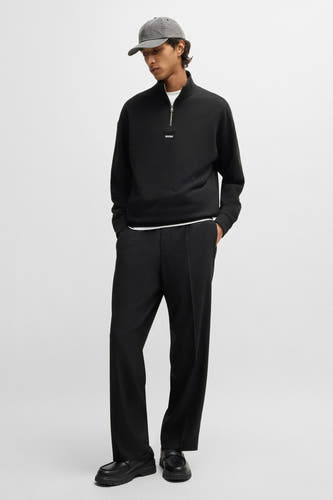 Pull-over noir avec fermeture à glissière de HUGO, porté avec un T-shirt blanc, un pantalon noir et une casquette grise.