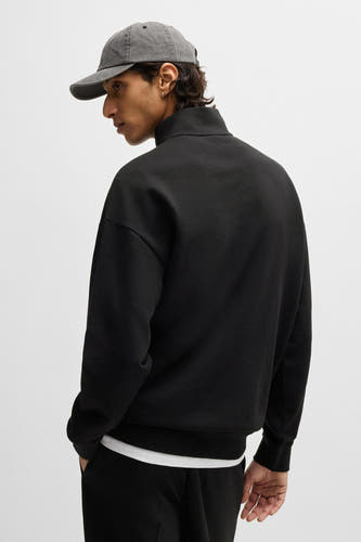 Pull noir HUGO, avec un col haut et des manches longues, porté avec un T-shirt blanc et un pantalon noir.