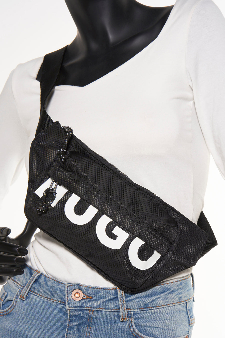 Zwarte schoudertas van HUGO, met een ruitjespatroon en witte merkletters, gedragen over een witte top en een jeans.
