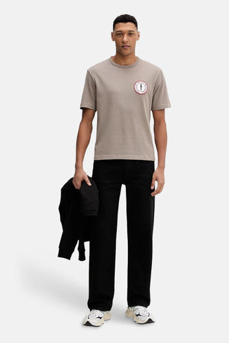 Bruin T-shirt van HUGO, met een ronde badge op de borst en gedragen met zwarte broek en sneakers.