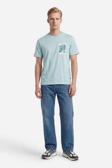 Turquoise T-shirt met korte mouwen van HUGO BOSS ORANGE, met palmbladprint en '7.8' op de borst, gedragen met blauwe jeans en sneakers.
