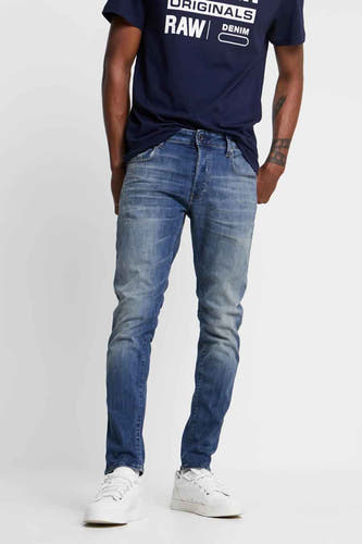 Slim fit jeans van G-Star RAW in middenblauw denim, met een donkerblauw T-shirt en witte sneakers.
