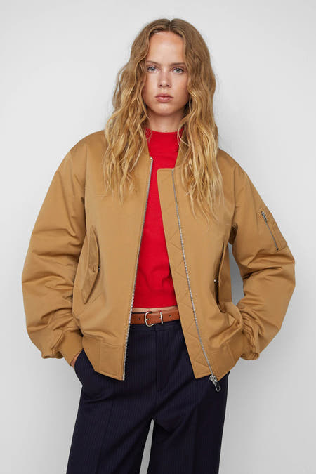 Bruine korte jas van MANGO, met lange mouwen, een rits en zakken, gedragen met een rode top en donkerblauwe broek.
