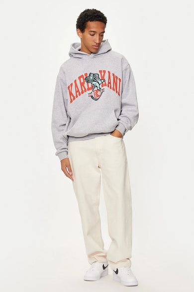 Grijze sweater met kap van Karl Kani, met een logo op de borst, gecombineerd met een lichte broek en witte sneakers.
