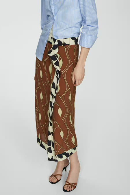 Bruine rok van MANGO, met een opvallend golvend patroon en zwart-witte details aan de zijkant, gecombineerd met een gestreept blauw overhemd.
