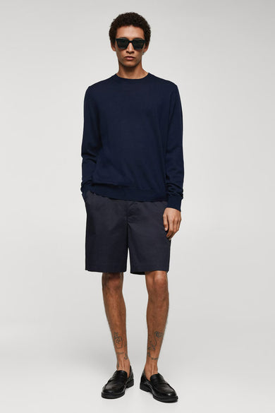 Blauwe short van MANGO, gedragen met een donkerblauwe trui en zwarte leren loafers, staand.
