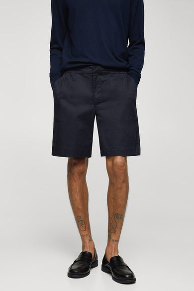 Blauwe short van MANGO, gedragen met een donkerblauwe trui en zwarte loafers, met tatoeages op de enkels.
