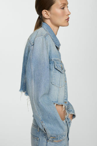 Denim jeansjas in middenblauw van MANGO, met een verkorte pasvorm en gerafelde details, in zij-aanzicht.
