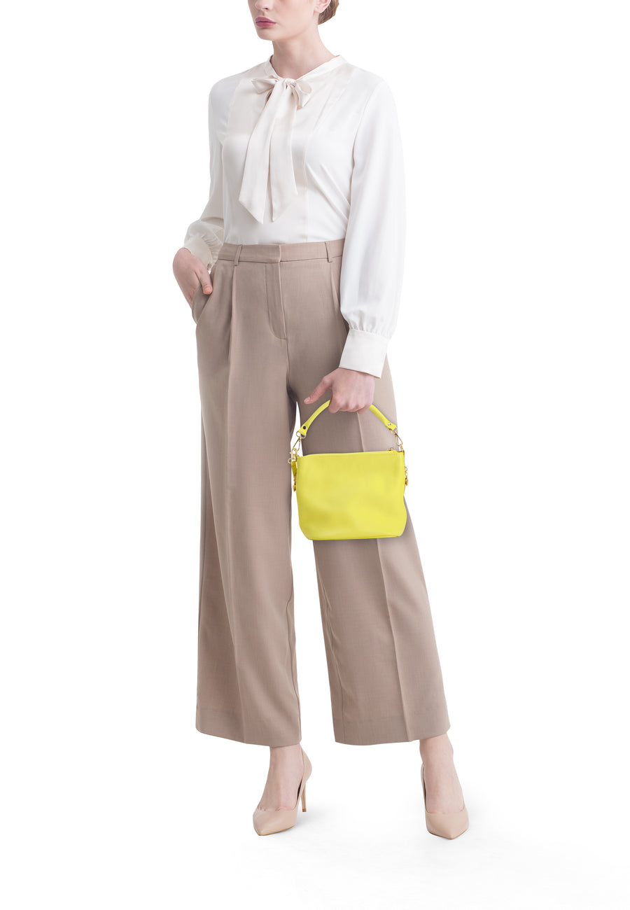 Gele schoudertas van Access®, met korrelige textuur en gouden hardware, gedragen door een vrouw in beige broek.
