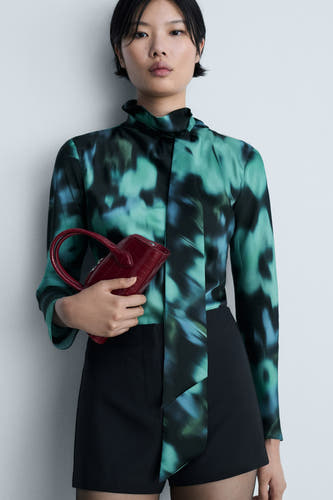 Multikleurige blouse met lange mouwen van MANGO, met een abstract patroon en gedragen met zwarte shorts en een rode handtas.
