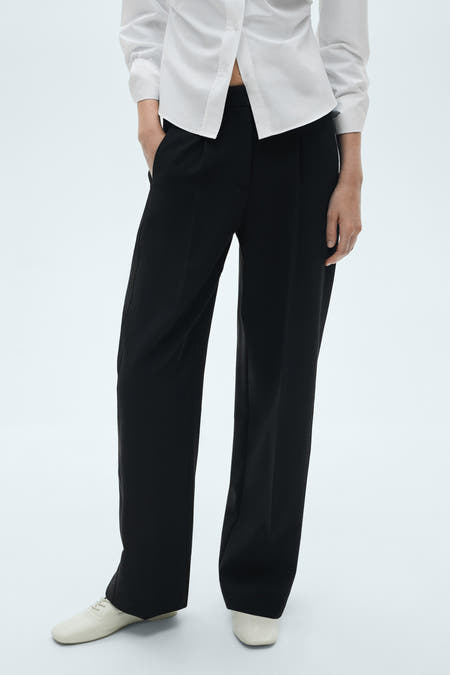 Zwarte broek van MANGO, met een wijde pijp en steekzakken, gecombineerd met een wit overhemd en platte schoenen.

