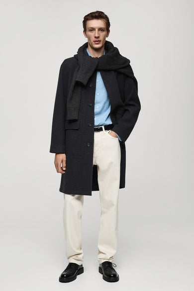 Manteau noir long de MANGO, avec un foulard, une chemise bleue et des jeans blancs.