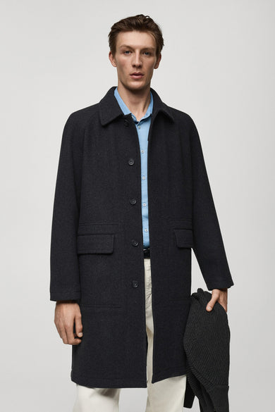 Long manteau noir de MANGO porté avec un chemisier bleu et un pantalon blanc.