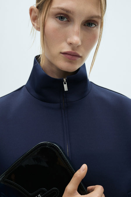 Blauw gilet van MANGO, met een hoge kraag en een zwarte tas in de hand.

