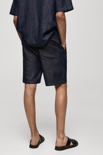 Jeansshort van mid blue denim van MANGO, achterkant, gecombineerd met een denim overhemd en zwarte sandalen.