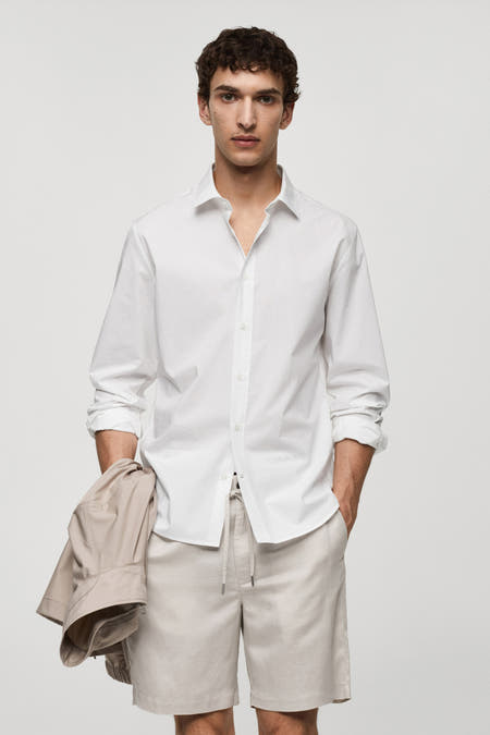 Blouse blanche à manches longues de MANGO, avec des manches retroussées, associée à des shorts légers et un manteau.
