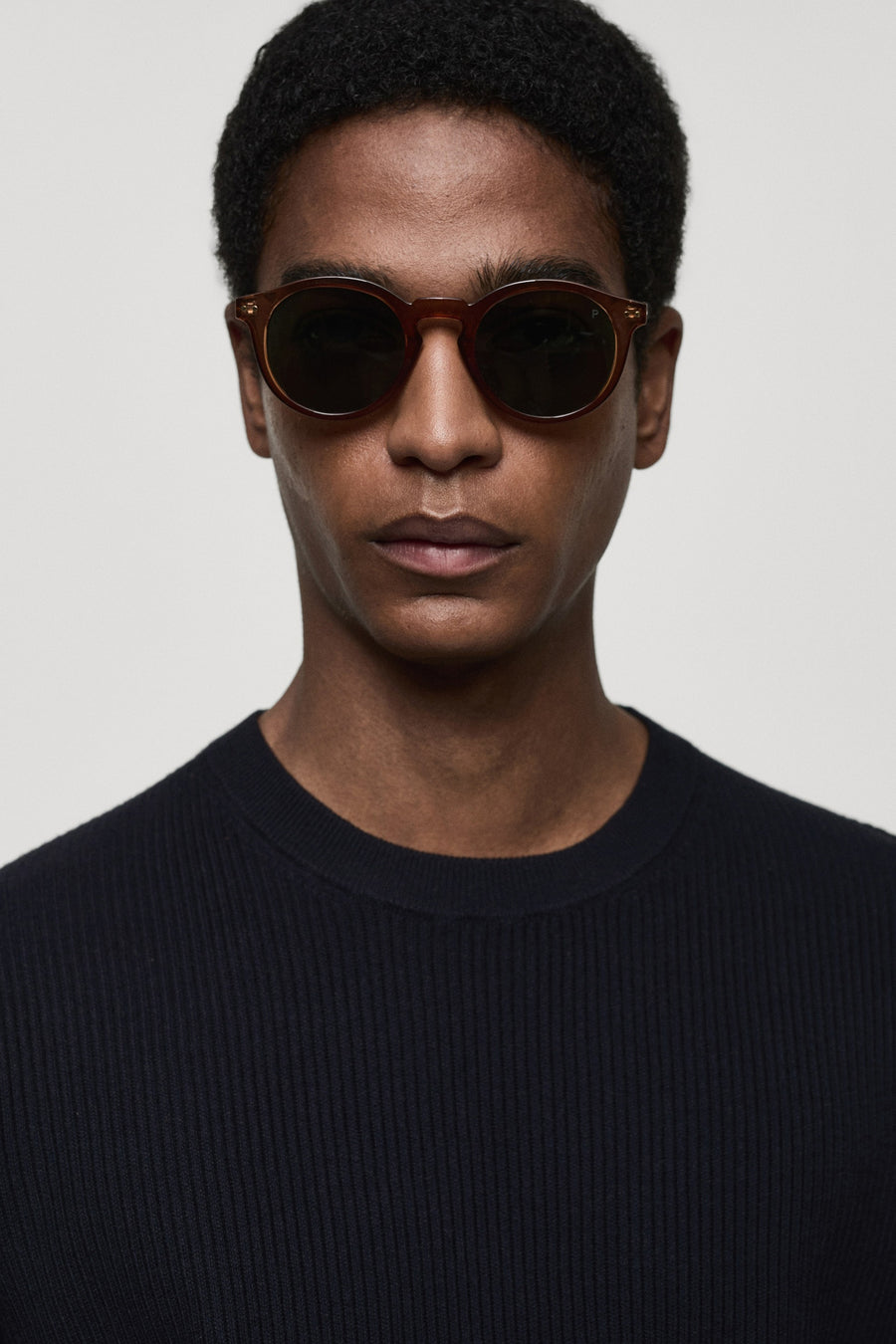 T-shirt bleu avec col rond de MANGO, porté avec un pull rayé foncé et des lunettes de soleil marron.