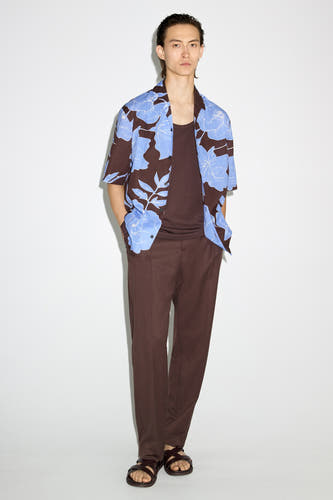Bruin overhemd met korte mouwen van MANGO, met een blauw bloemenpatroon, gedragen over een bruin hemd en een bruine pantalon met sandalen.
