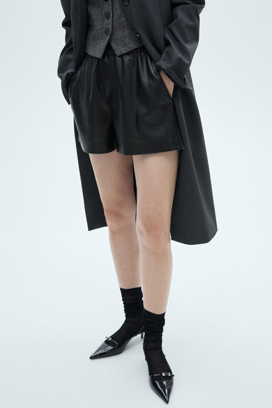 Zwarte bermuda van MANGO, gecombineerd met een grijs vest en een lange jas, gedragen met zwarte sokken en puntschoenen.
