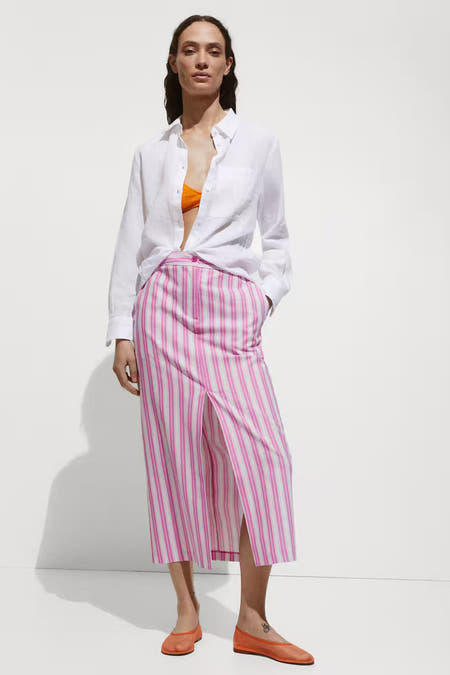 Lange roze gestreepte rok van MANGO, met een split aan de voorkant, gecombineerd met een wit overhemd en oranje schoenen.
