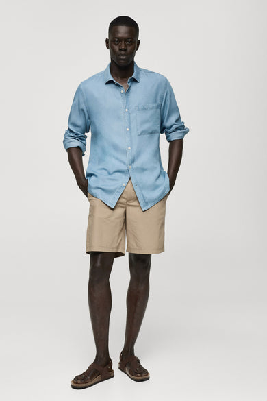 Jeanshemd van midblauw denim van MANGO, met lange mouwen opgerold en beige shorts met sandalen.
