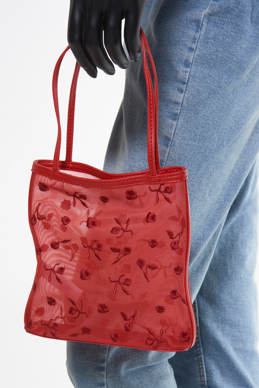 Rode handtas van MANGO, met bloemenborduurwerk, gehouden door een zwarte mannequinhand, met een spijkerbroek op de achtergrond.