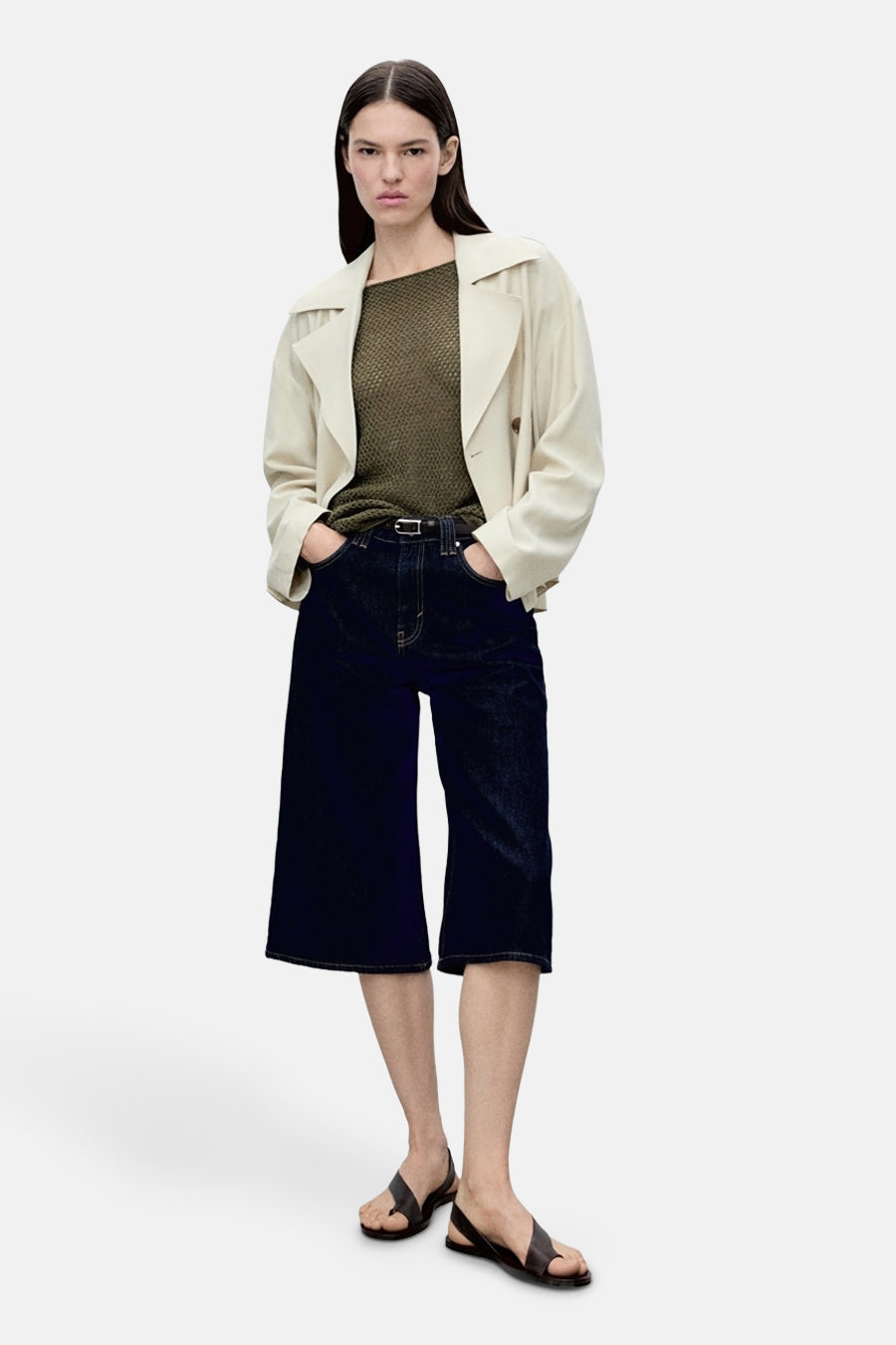 Vrouw met lichte trenchcoat, olijfgroene top, donkerblauwe culottes en leren sandalen.
