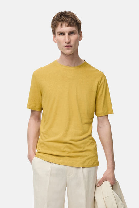 Geel T-shirt met korte mouwen van MANGO, gedragen met een crèmekleurige broek, met een jas in de hand.