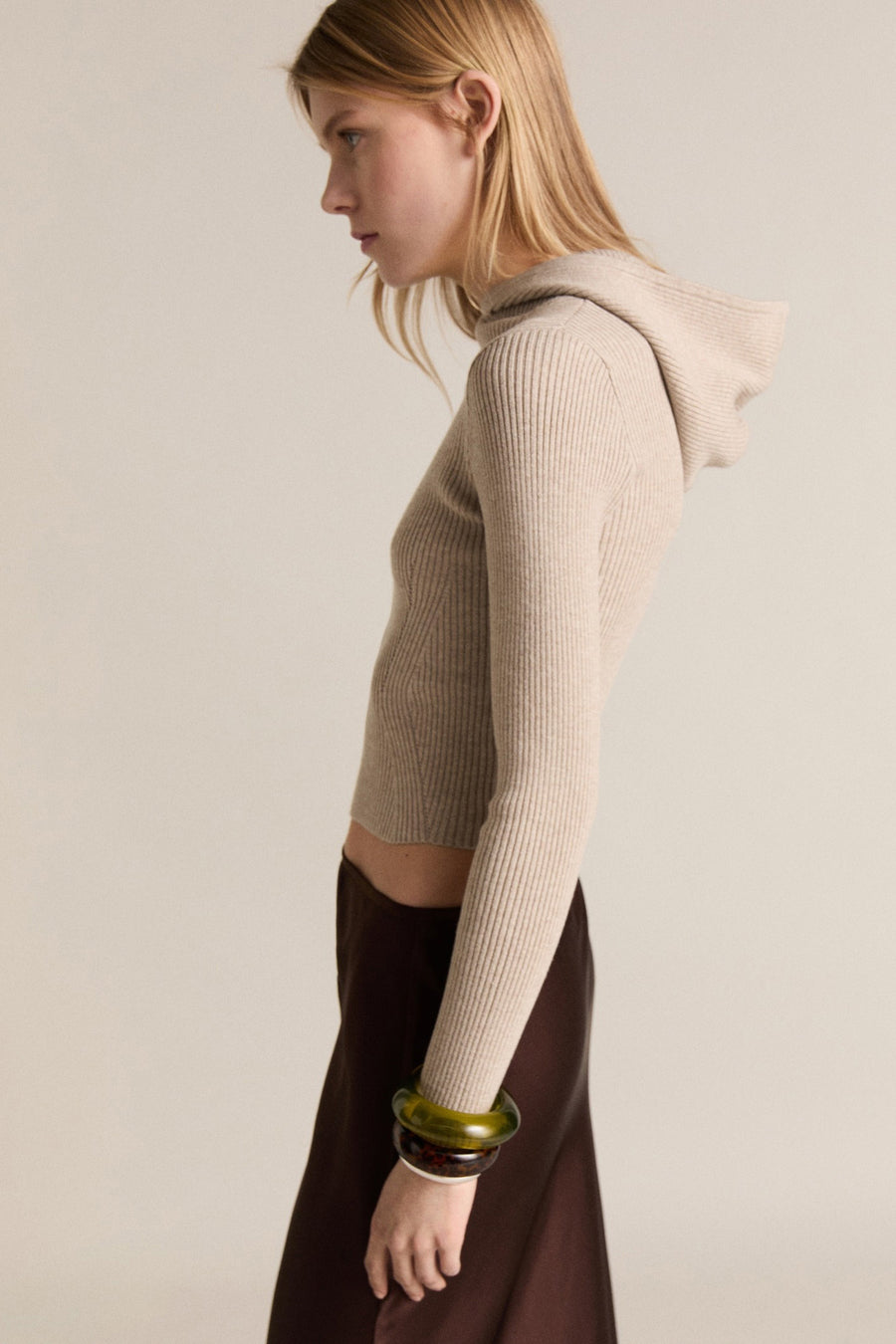 Beige trui met ronde hals van MANGO, geribd en met lange mouwen, gedragen met een bruine rok en groene armbanden.
