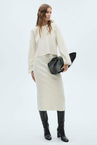 Pull-over blanc avec col rond de MANGO, porté avec une jupe et des bottes noires et un sac.