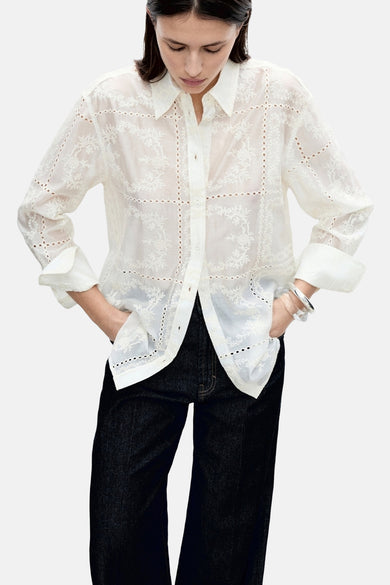 Vrouw in een witte broderie blouse en zwarte jeans, staand met handen in de zakken.