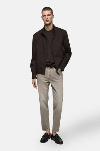 Grijze tapered jeans van MANGO, gecombineerd met een donkerbruin jack en zwarte leren loafers.