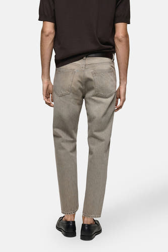 Grijze tapered jeans van MANGO, gezien van achteren, met een donkerbruin shirt en zwarte schoenen.
