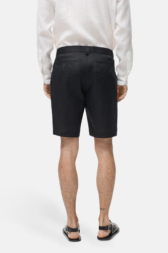Zwarte short van MANGO, achterkant zichtbaar, gecombineerd met een wit linnen overhemd en zwarte sandalen.