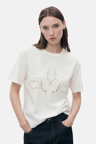 Wit T-shirt met korte mouwen van MANGO, met een sierlijk opengewerkt detail op de voorkant, gecombineerd met een donkere jeans.
