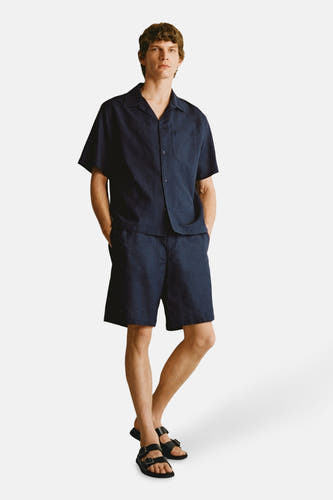 Chemise bleue à manches courtes de MANGO, avec col boutonné et poche brodée, associée à des shorts bleu marine et des sandales.