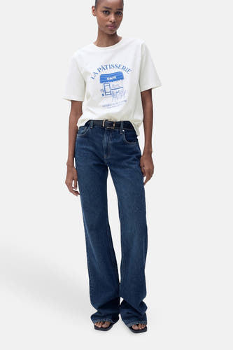 T-shirt blanc à manches courtes de MANGO, avec une impression bleue d'un café, porté avec des jeans foncés et des sandales.