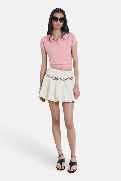 Polo rose à manches courtes de MANGO, porté avec une mini-jupe blanche et une ceinture en chaîne argentée, des sandales noires.