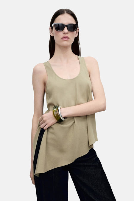 Mouwloze blouse in kaki, van MANGO, met een asymmetrische zoomlijn, gedragen met een jeans en zonnebril.
