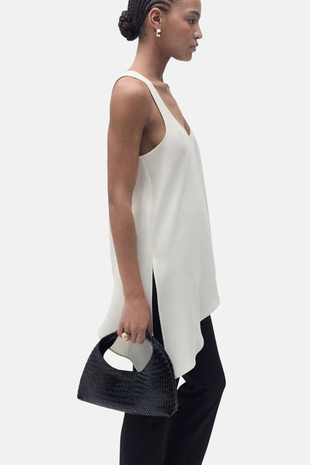 Mouwenloze ecru blouse van MANGO, met V-hals en zijsplit, gecombineerd met zwarte pantalon en een zwarte gevlochten handtas.