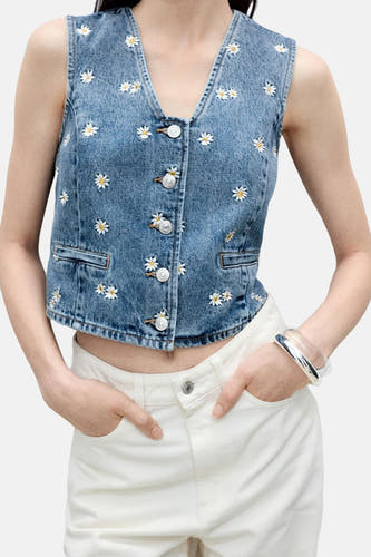 Mouwloze denim blazer in middenblauw van MANGO, met margrietjespatroon en witte broek en zilveren armband.

