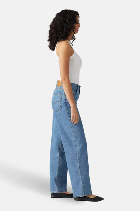 Vrouw in witte tanktop en wijde, blauwe spijkerbroek, staand.