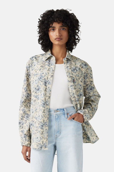 Vrouw met gekruld haar, bloemenoverhemd en spijkerbroek.