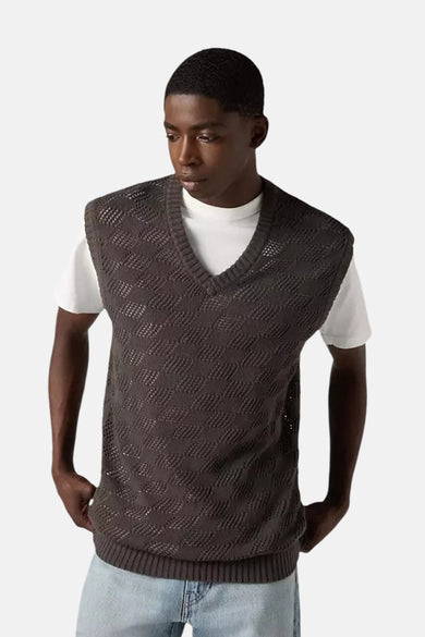 Man in grijs gebreid vest met ruitpatroon, witte T-shirt en jeans tegen witte achtergrond.
