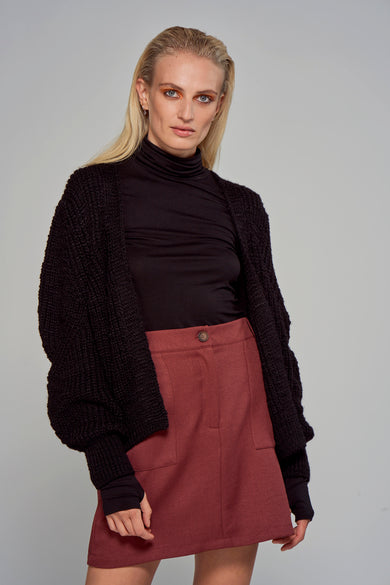 Zwart gilet van Astrid Black Label, met een open voorkant en gebreide textuur, over een zwarte coltrui en een rode rok.
