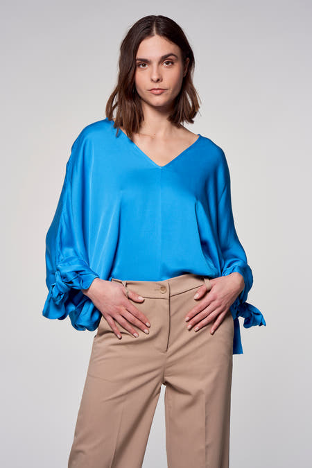 Blauwe blouse van Astrid Black Label met lange mouwen en strikdetails, gecombineerd met beige pantalon.
