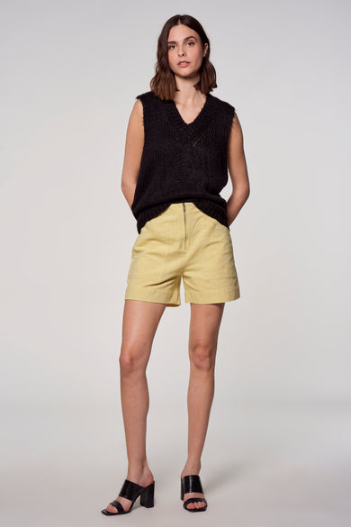 Gele short van Astrid Black Label, met hoge taille en zichtbare ritssluiting, gecombineerd met zwarte top en hakken.
