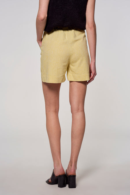 Gele short van Astrid Black Label, met een lichte textuur, gedragen met zwarte top en hoge hakken, gezien van achteren.

