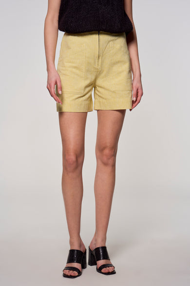 Gele short van Astrid Black Label, met steekzakken en een ritssluiting, gecombineerd met zwarte top en hakken.
